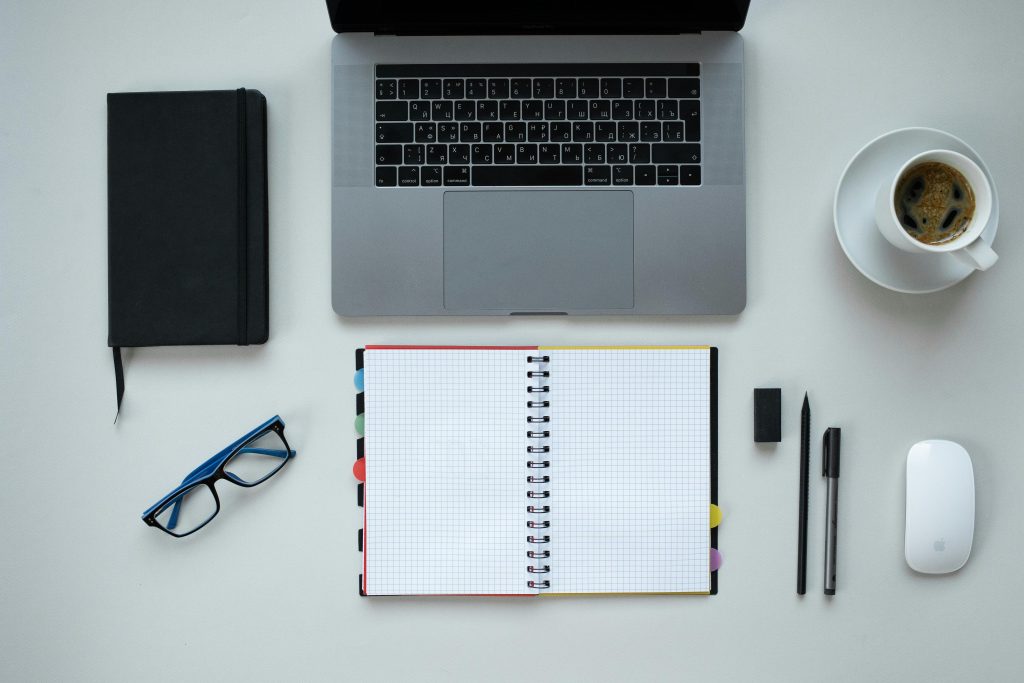 A pair of glasses, a black diary,An open diary,two pens, a cup of coffee, a white wireless mouse and a laptop