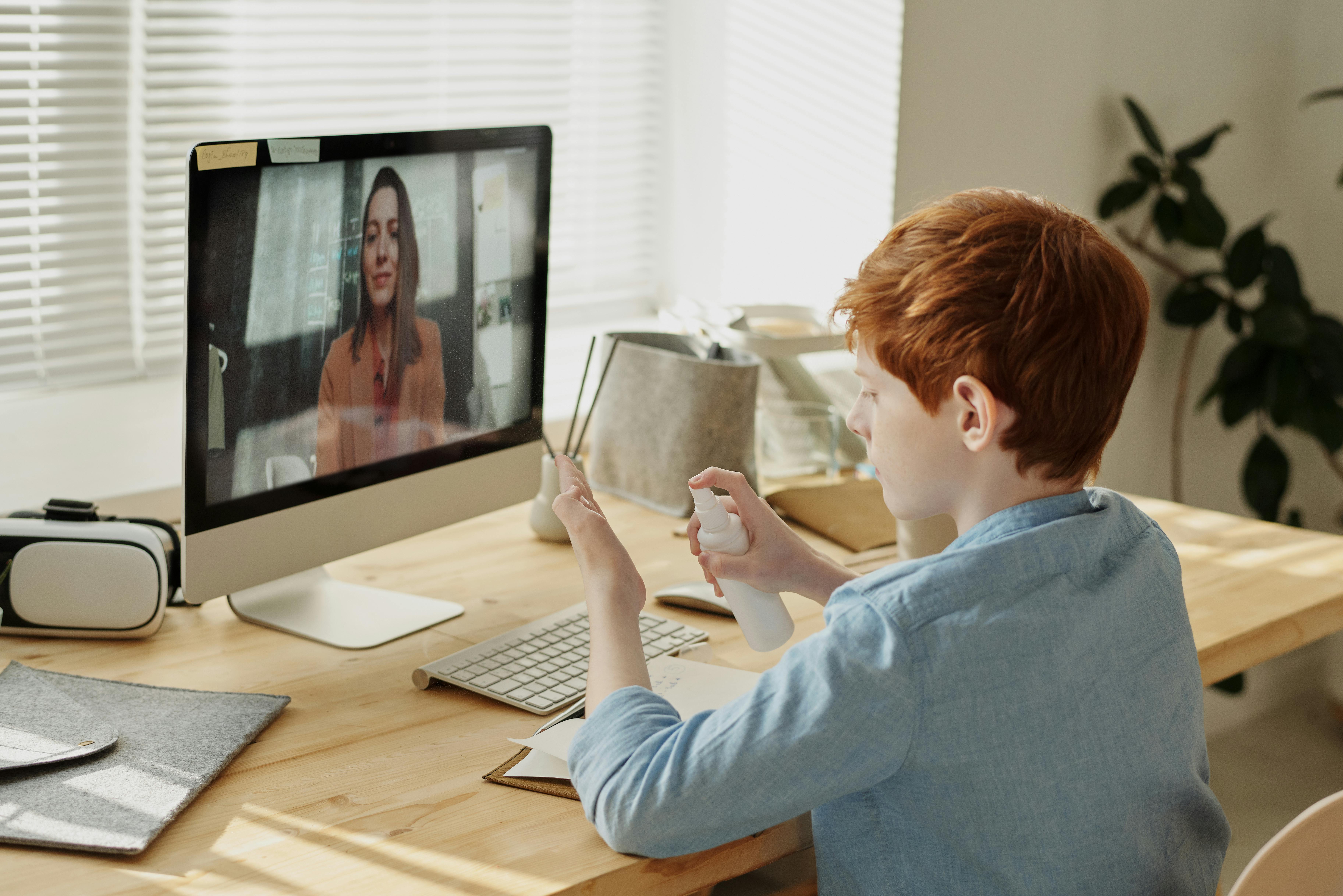 A young ginger haired boy seated at a desk in front of a computer that has a picture of a young brunette caucasion woman.