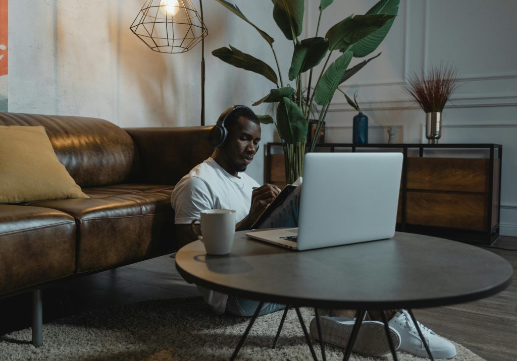 Black French man seated on the ground with headphones on a laptop in front of him and a cup of coffee besides him